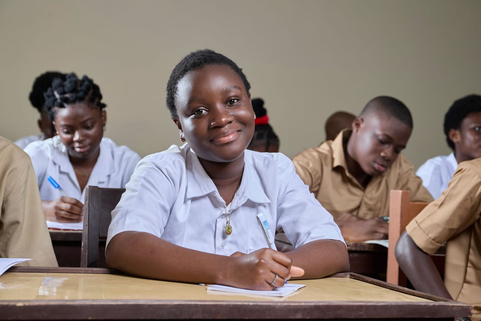 A secondary school student smiling at her desk, pen in hand, classmates working behind her.