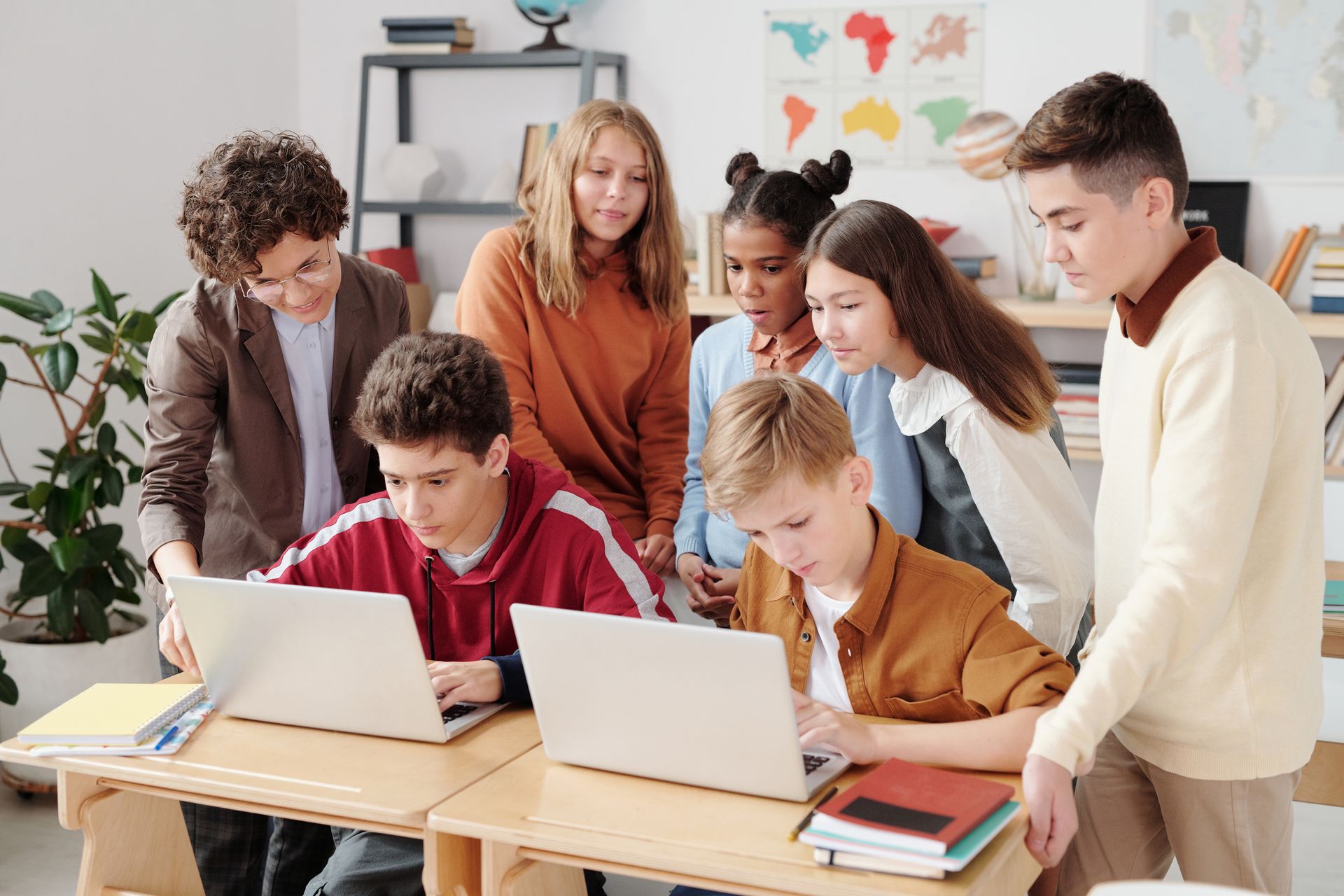Teenagers collaborating on laptops in a classroom, guided by a teacher.